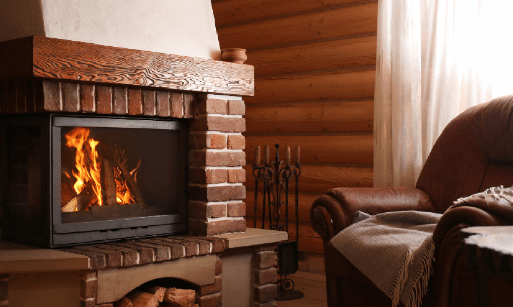 A cozy living room with a lit fireplace surrounded by brick and wood accents. A plush armchair draped with a blanket is positioned nearby. A side table holds two glasses of wine, creating a warm and inviting atmosphere.