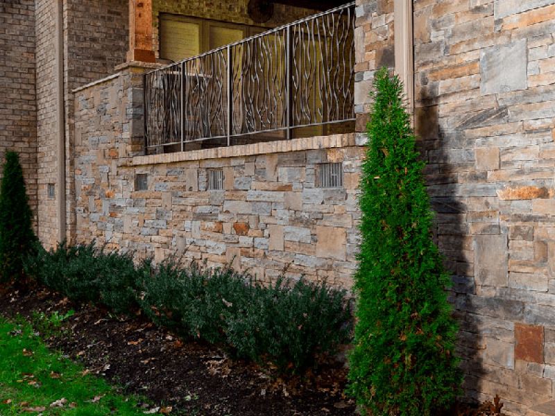 A stone building facade with a decorative metal railing on a balcony. Below, there is a garden with green shrubs and a conical evergreen tree planted in mulch, bordering a grass lawn.