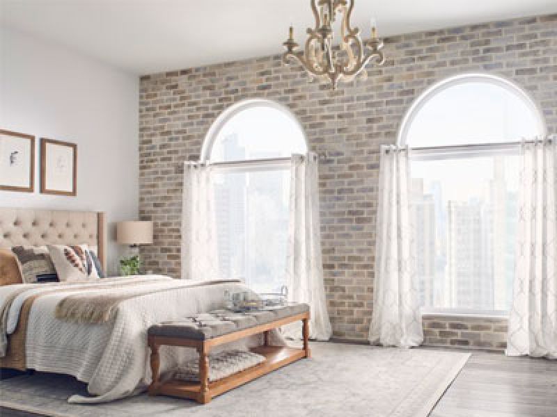 A cozy bedroom with a brick accent wall, featuring large arched windows with sheer curtains. The room includes a plush bed with neutral linens, a bench at the foot, a chandelier, and framed art on the wall. Natural light brightens the space.