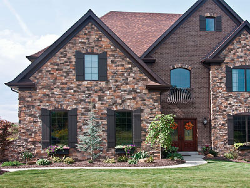 A large brick house with a gabled roof and several windows. The front yard has a well-maintained lawn and landscaped plants. The entrance features a double door with fall wreaths, and a curved walkway leads to the steps.