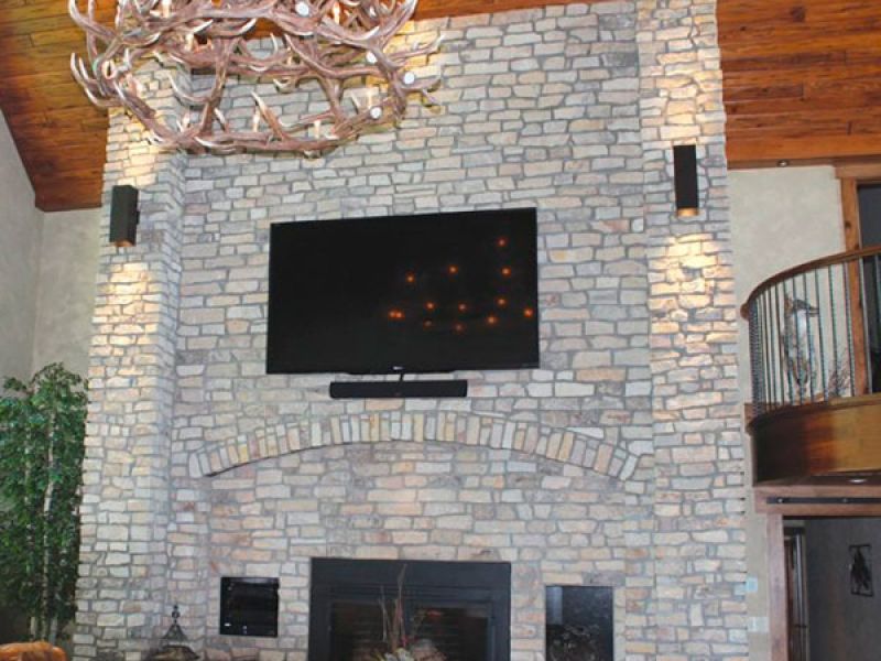 A cozy living room with a tall stone fireplace, mounted TV, and antler chandelier. Leather chairs surround a circular coffee table. A balcony with a wooden railing is visible in the background.
