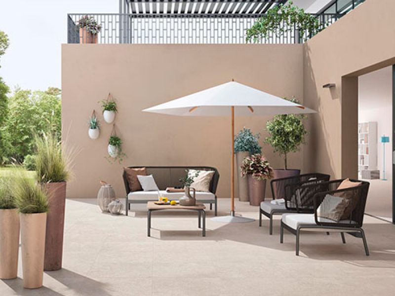 A modern outdoor patio with a beige wall, featuring a seating area with black chairs, a white umbrella, potted plants, and a table set with a pitcher and glasses. Decorative wall planters add greenery, while trees are visible in the background.