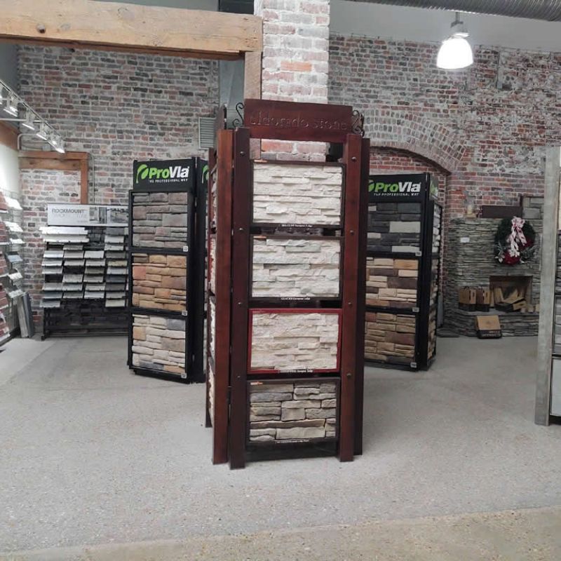 A showroom with displays of stone veneer samples. The central display features various stacked stone styles. Shelves on the left hold additional samples, and the brick walls and industrial lighting enhance the rustic atmosphere.