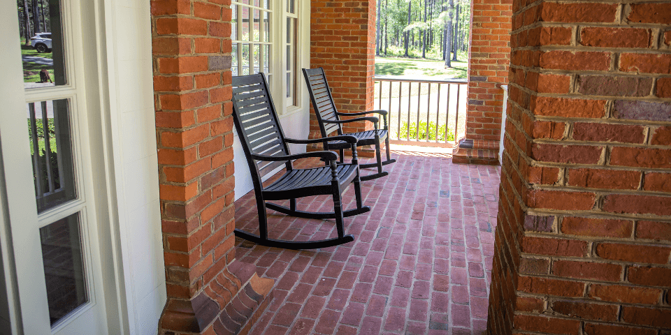 Two black rocking chairs sit on a brick porch, surrounded by red brick walls and overlooking a lawn with trees in the background.