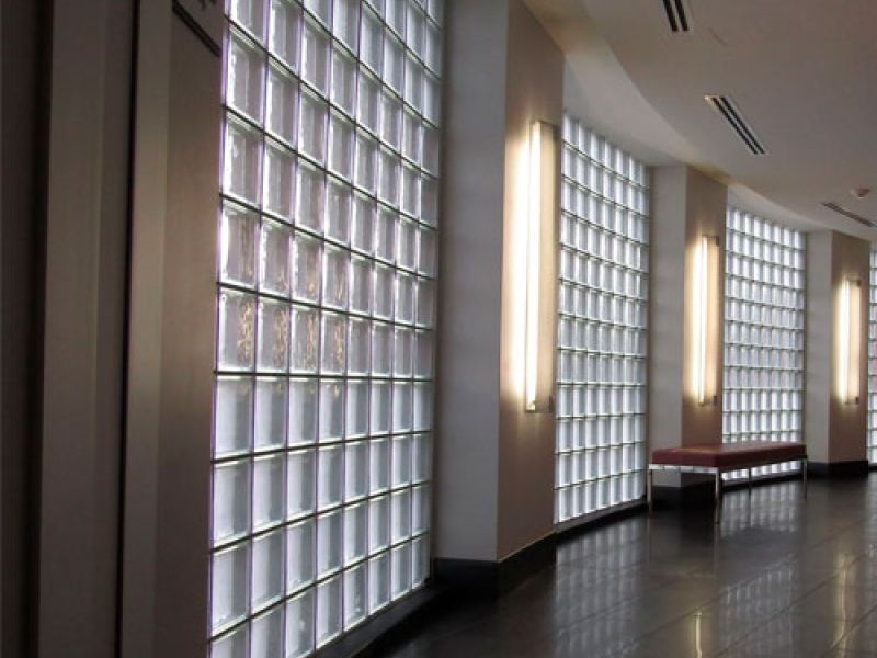 A modern hallway with large glass block windows on one side, illuminating the space with natural light. The floor is dark and glossy, and there are benches placed along the windowed wall. Ceiling lights complement the bright atmosphere.