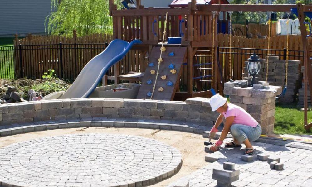 A person arranges bricks in a circular pattern on a patio, beside a playground with a slide, climbing wall, and swing set, surrounded by a fenced backyard.