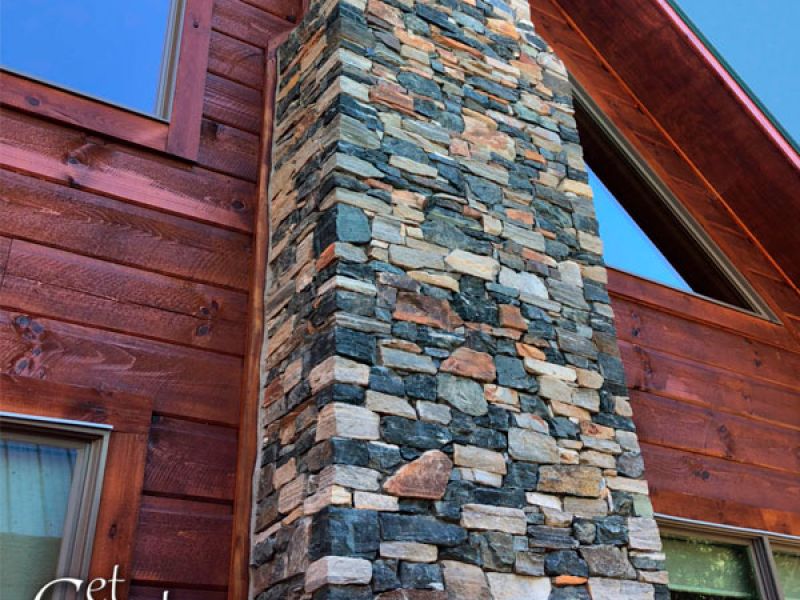 A rustic wooden house with a tall, stone chimney is shown against a clear blue sky. The stone chimney features a mix of gray, brown, and beige stones. The house has large triangular windows, and part of the logo Get Real Stone is visible.
