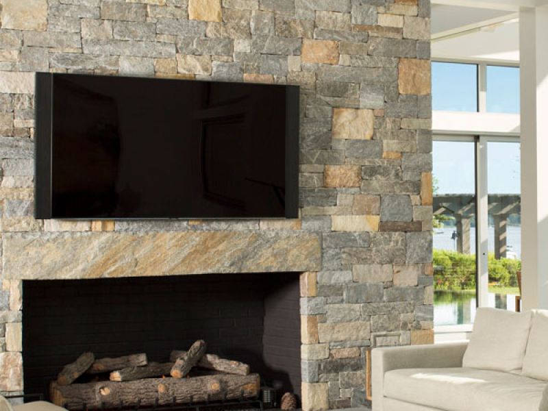 A modern living room features a stone accent wall with a large flat-screen TV mounted above a fireplace filled with stacked logs. Natural light pours in through large windows, illuminating a beige sofa and sleek furniture.