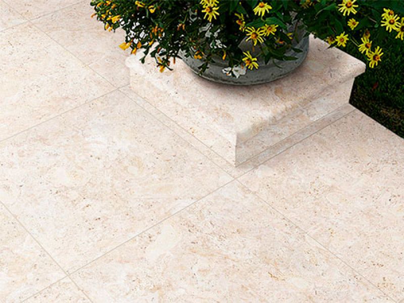 A close-up of light beige stone tiles on a patio. A planter filled with yellow flowers with green leaves sits on the tiles, partially visible.