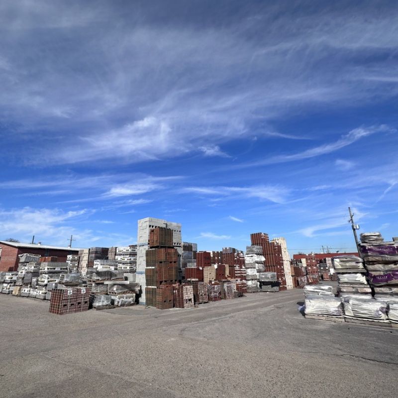 A wide view of a construction materials yard with stacks of bricks, concrete blocks, and pallets wrapped in plastic under a blue sky with scattered clouds. Several industrial buildings are visible in the background.