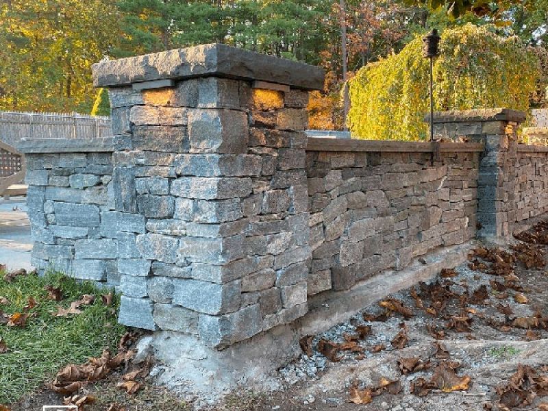 A sturdy stone wall with decorative pillars stands amidst fallen autumn leaves. The warm glow of light highlights the texture of the stone, while trees with green and yellow foliage are visible in the background.