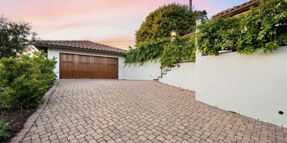 A wooden garage door sits at the end of a brick driveway, flanked by green foliage and a white wall, at dusk. Clean and well-placed brick pavers make up wide driveway