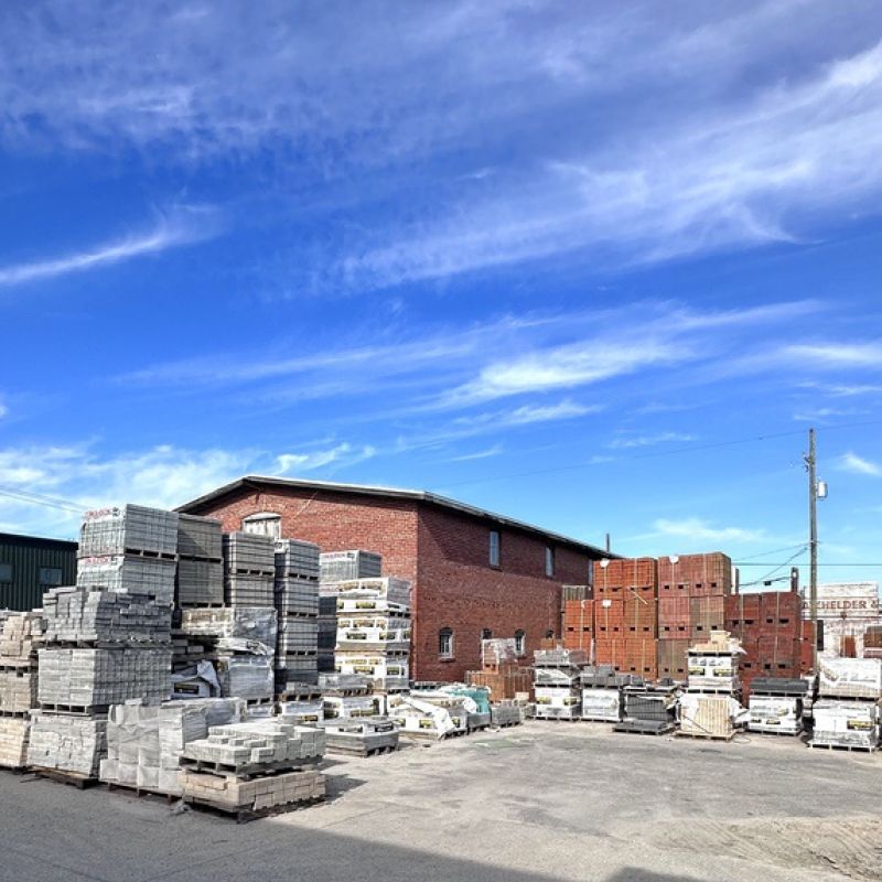 A construction supply yard with stacks of bricks and concrete blocks under a bright blue sky with wispy clouds. A brick building in the background and signs of industrial activity are visible.
