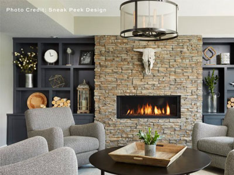 A cozy living room with a stone fireplace at the center, a mounted skull above, and a modern chandelier. Two gray armchairs face the fire. A wooden tray with decorations sits on the table. Shelving with decor and books frames the scene.