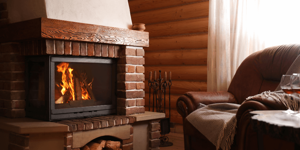 A cozy living room with a lit fireplace surrounded by brick and wood accents. A plush armchair draped with a blanket is positioned nearby. A side table holds two glasses of wine, creating a warm and inviting atmosphere.