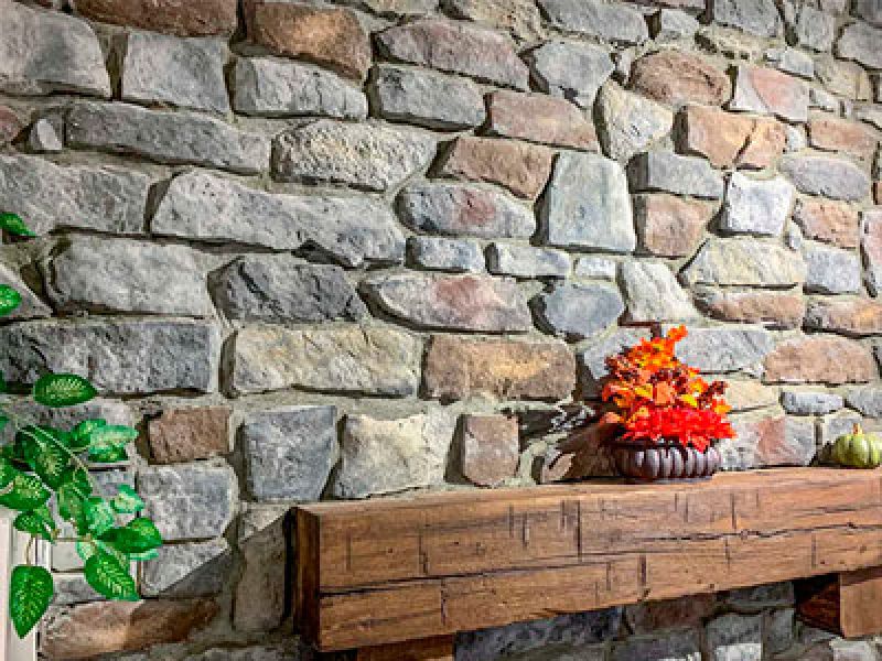 A rustic stone wall with a wooden mantel. On the mantel, there is a small pot with red and orange autumn leaves and a decorative pumpkin. A green leafy plant is partially visible on the left side.