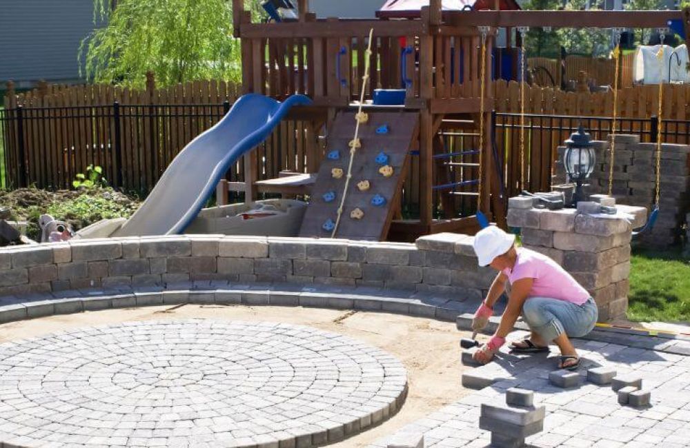 A person arranges bricks in a circular pattern on a patio, beside a playground with a slide, climbing wall, and swing set, surrounded by a fenced backyard.