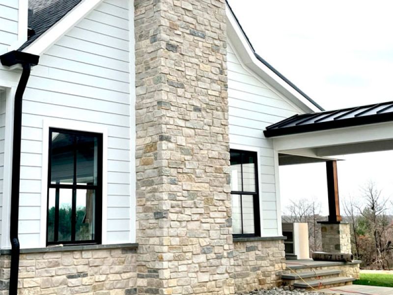 A modern house exterior featuring white siding and a stone chimney. Black-trimmed windows complement the design, and the entrance has a small porch with stone steps. The surrounding landscape includes grass and a rocky path.