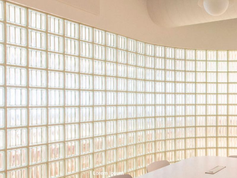 A modern conference room with a curved wall made of frosted glass blocks allowing soft light to enter. A long, white table with several chairs surrounds the room. Minimalist ceiling lamps hang above.