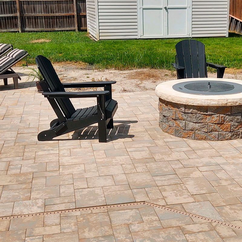 A sunny outdoor patio with two black Adirondack chairs next to a stone fire pit on a patterned stone surface. A neatly maintained lawn and a white shed are in the background.