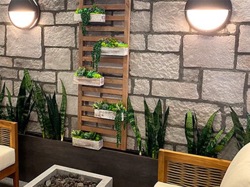 A cozy sitting area with beige chairs and a stone wall backdrop. A vertical wooden panel features hanging planters with green foliage. Snake plants line the bottom, and two wall lights add a warm ambiance. A rectangular stone planter is in front.