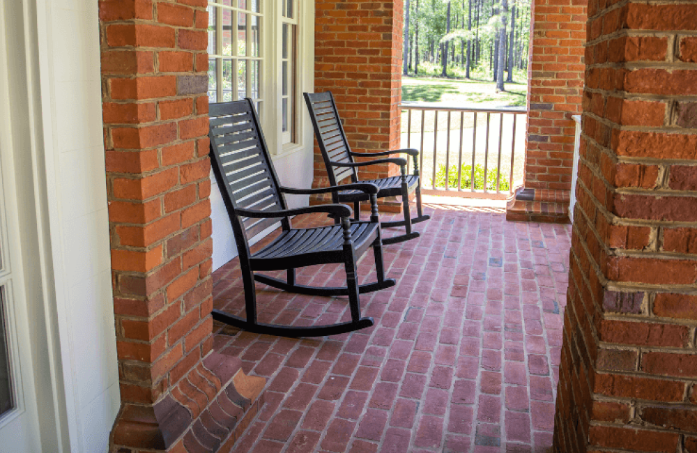 Two black rocking chairs sit on a brick porch, surrounded by red brick walls and overlooking a lawn with trees in the background.