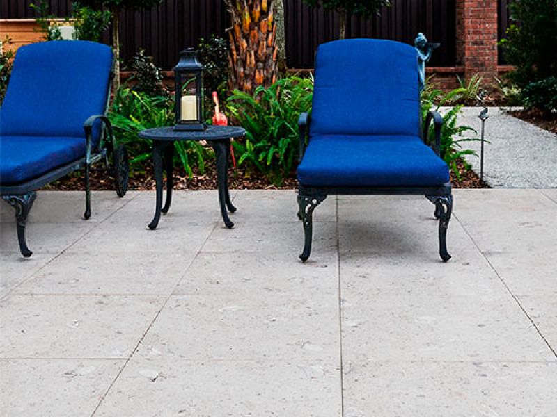 Two blue cushioned chairs with wrought iron frames are arranged around a small black metal table. A lantern and a small pepper decoration sit on the table. The patio has light stone flooring, and green plants are in the background.