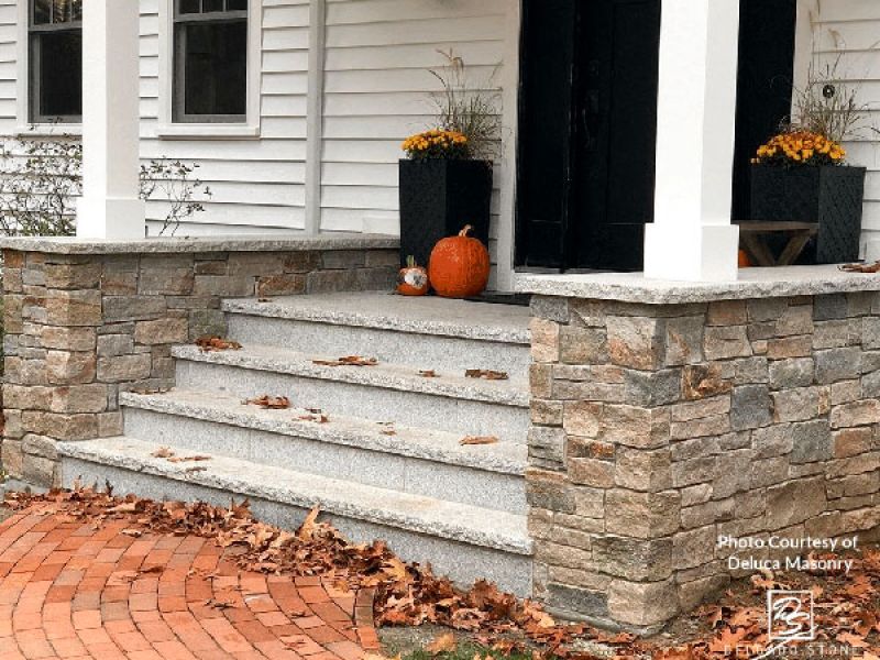 A stone porch with granite steps is adorned with autumn leaves, pumpkins, and potted flowers. The entrance features white siding and a black door, bordered by a curved brick pathway.