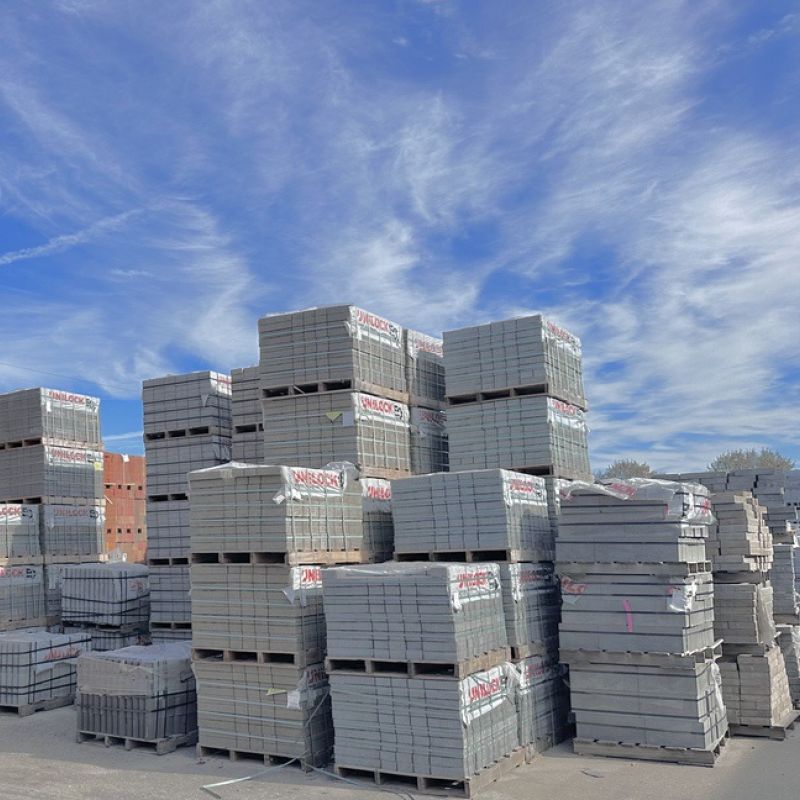 Stacks of gray bricks are arranged on wooden pallets in an outdoor storage area under a blue sky with scattered clouds.