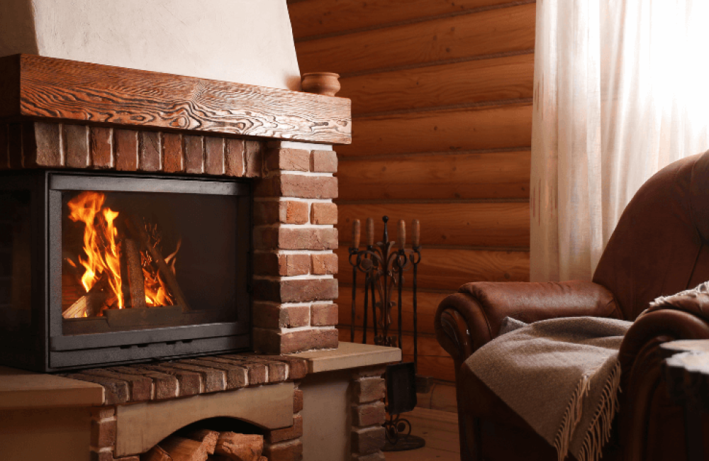 A cozy living room with a lit fireplace surrounded by brick and wood accents. A plush armchair draped with a blanket is positioned nearby. A side table holds two glasses of wine, creating a warm and inviting atmosphere.