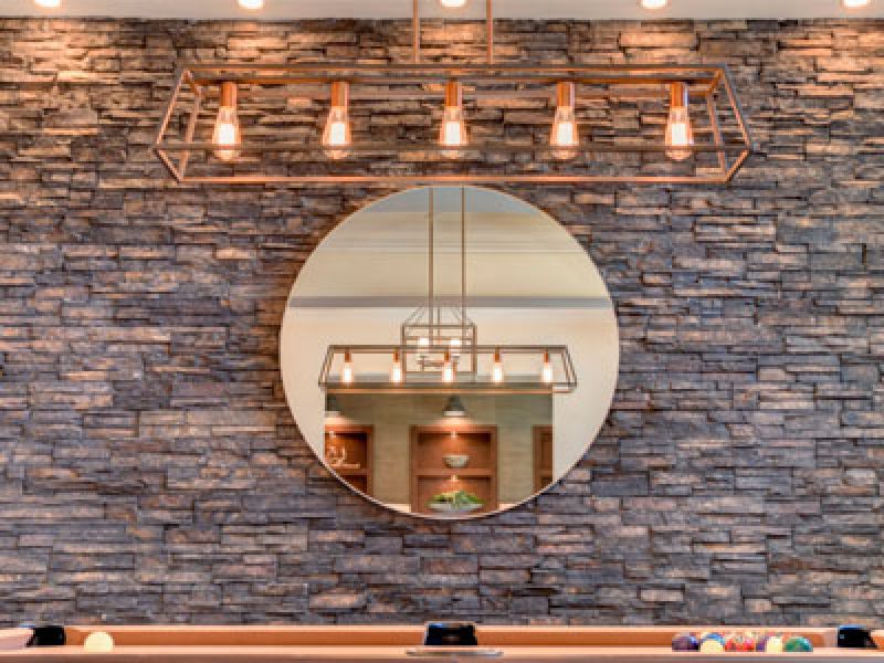 A cozy room with a circular mirror on a stone wall, reflecting stylish pendant lights. Below the mirror, theres a wooden pool table with colorful billiard balls. Warm lighting creates an inviting ambiance.
