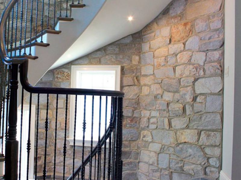 A spiral staircase with dark wooden railings curves upward beside a stone wall. A small window is visible, allowing light to illuminate the textured stones. The flooring is wooden, complementing the rustic style of the interior.