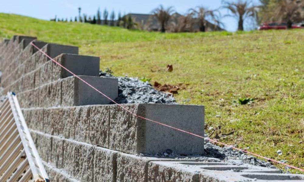 Concrete blocks form a retaining wall under construction, supported by a guiding string. They are set against a sloping grassy hill with trees visible in the background.