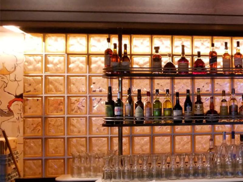 A bar counter with rows of glasses sits in front of a wall made of glass blocks. Shelves hold various bottles of alcohol. The wall is illuminated from behind, creating a warm glow. A mural is partially visible on the left side.