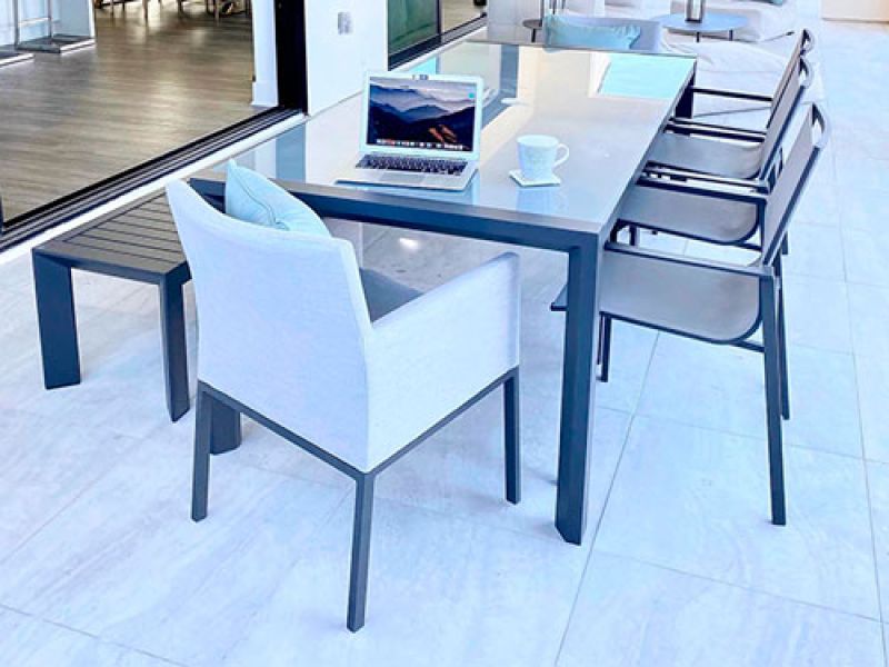 Modern outdoor patio setup with a sleek rectangular table surrounded by chairs. A laptop displaying a scenic mountain view and a coffee cup on a saucer are placed on the table. The area is bright with natural light streaming in.