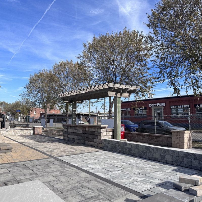 Outdoor area with stone pathways, a pergola, and brick structures. Trees line the scene under a clear sky with wispy clouds. A fence separates the area from a street with a building.