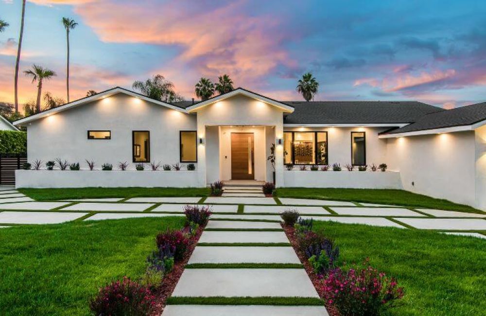 A modern single-story house stands at dusk, with geometric lawn patterns made of hardscapes and decorative shrubbery under a colorful sky.
