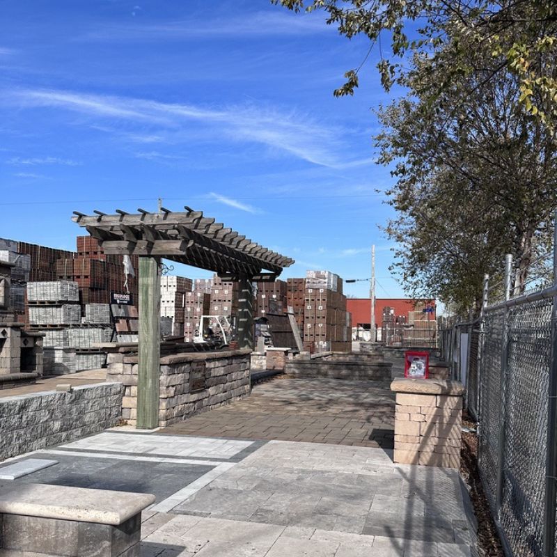A paved walkway with a wooden pergola and stone benches under a clear blue sky. Trees line the right side, and buildings and a fence are visible in the background.