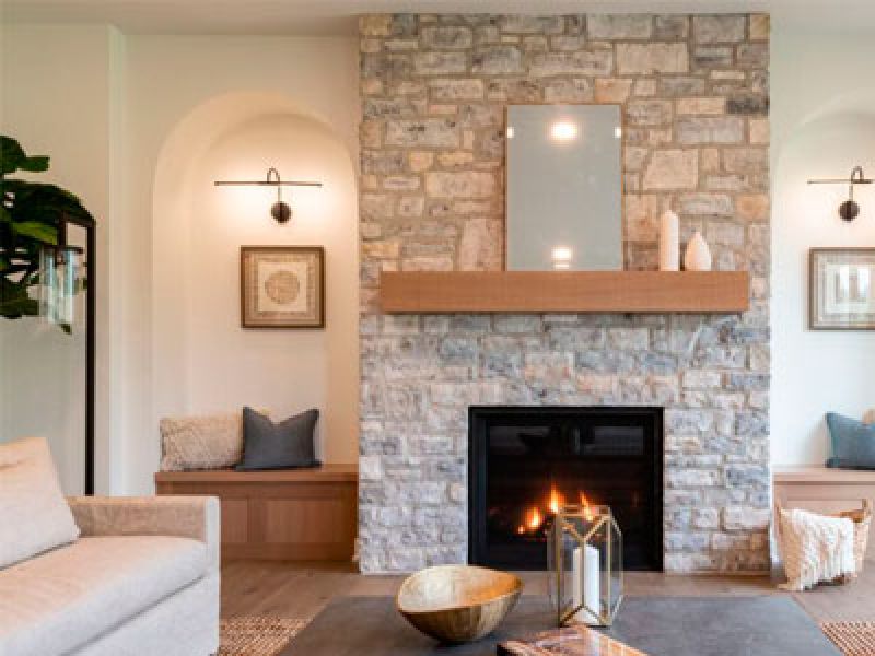 A cozy living room with a stone fireplace, two built-in benches with cushions on each side, and a mantle with decorative items. A comfortable sofa and a modern coffee table are in the foreground. Warm lighting creates an inviting atmosphere.