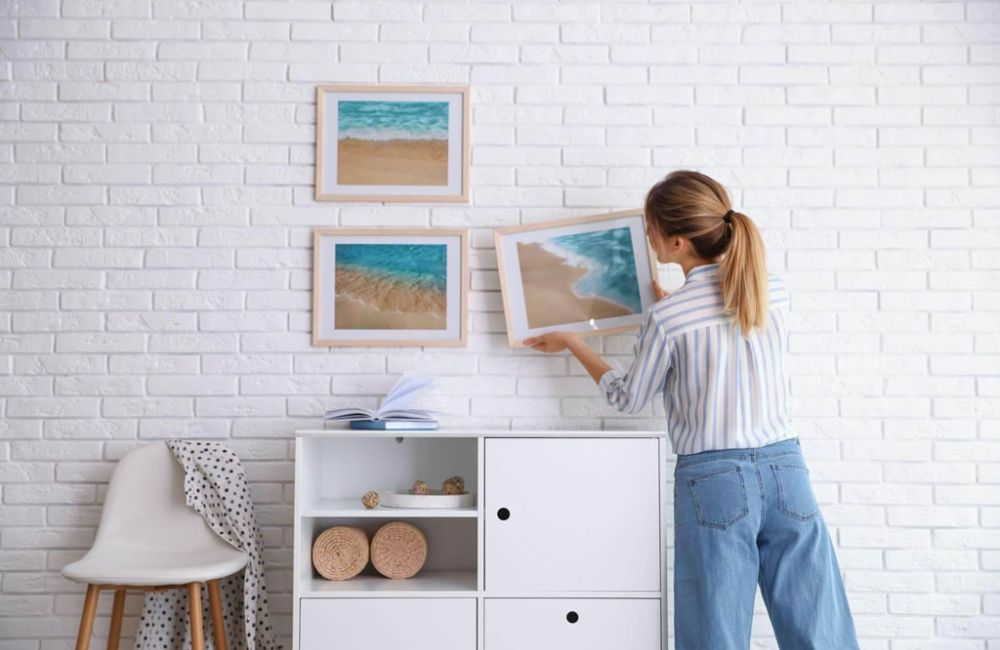 Woman arranging framed ocean artworks on a white brick wall for modern home decor.
