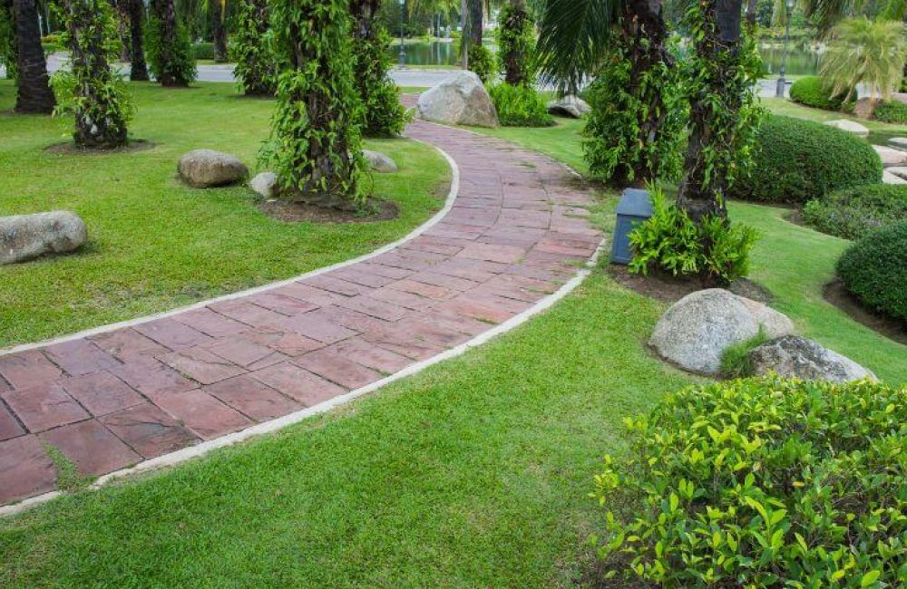 A winding red brick pathway meanders through a lush green park with manicured shrubs, palm trees, and interspersed large rocks.