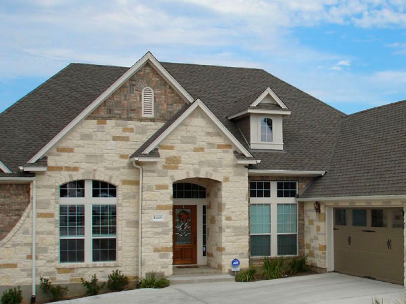 A modern suburban house with stone and brick exterior, featuring a prominent gabled roof. It has large windows, a wooden front door, and an attached two-car garage. A well-maintained driveway and minimal landscaping are visible under a partly cloudy sky.
