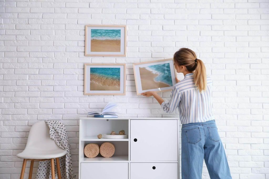 Woman arranging framed ocean artworks on a white brick wall for modern home decor.