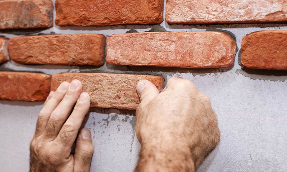 Hands installing red brick veneer on a wall for home construction or renovation.