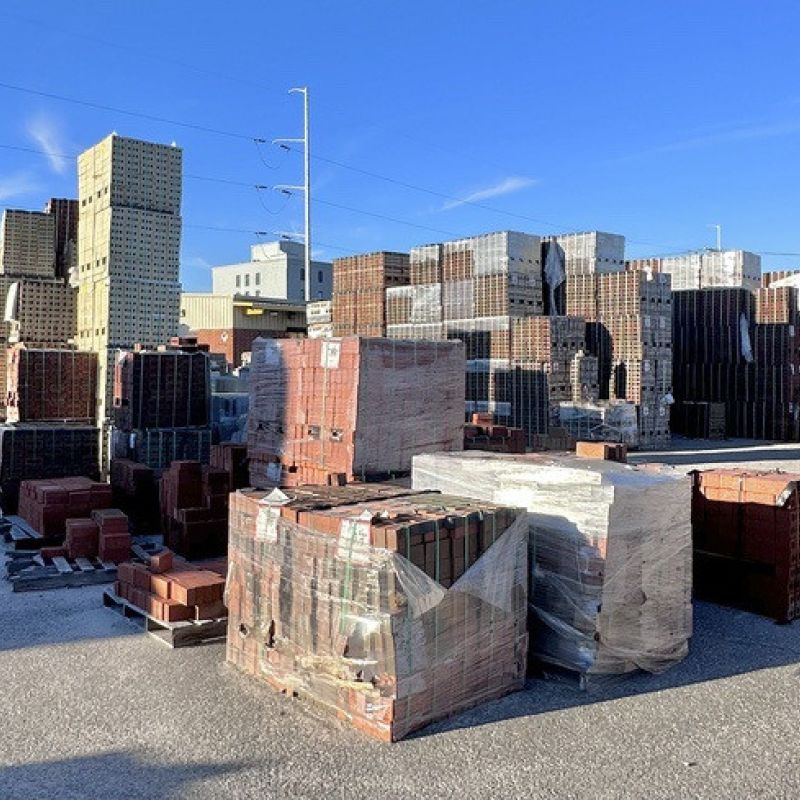Stacks of various types of bricks and construction materials are arranged neatly in a wide outdoor storage area. The clear blue sky and a few buildings are visible in the background.