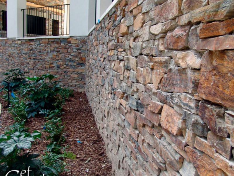 Close-up of a textured stone wall with earthy tones, surrounded by lush green plants. A building with a balcony and black railing is visible in the background. The image includes the text Get Real Stone.