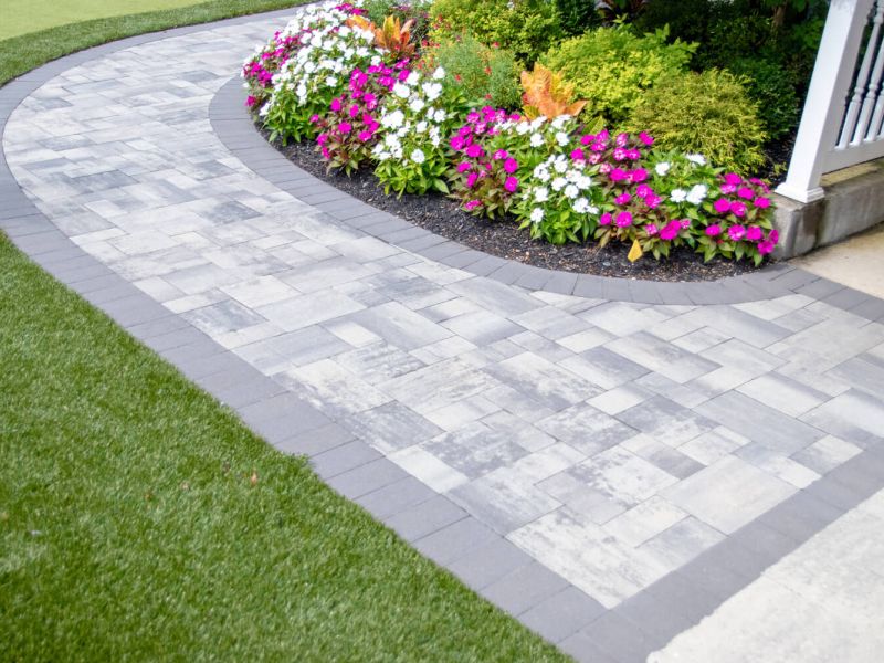 Curved stone paver walkway with vibrant flower bed and green lawn in residential garden setting.