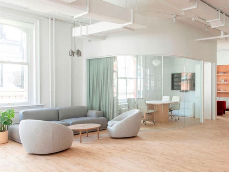 A modern office space with light wood flooring and white walls. It features a seating area with a gray sofa and two chairs, a circular coffee table, and a glass-walled conference room with a table, chairs, and a TV. Large windows let in natural light.