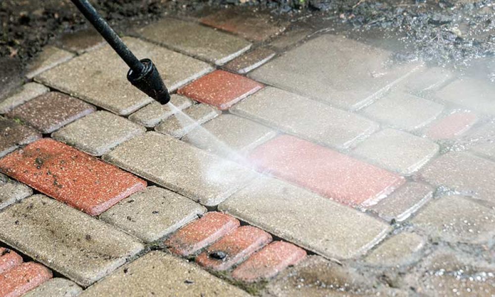 Pressure washing red and beige brick patio, removing dirt and grime with high-pressure water jet for a clean surface.