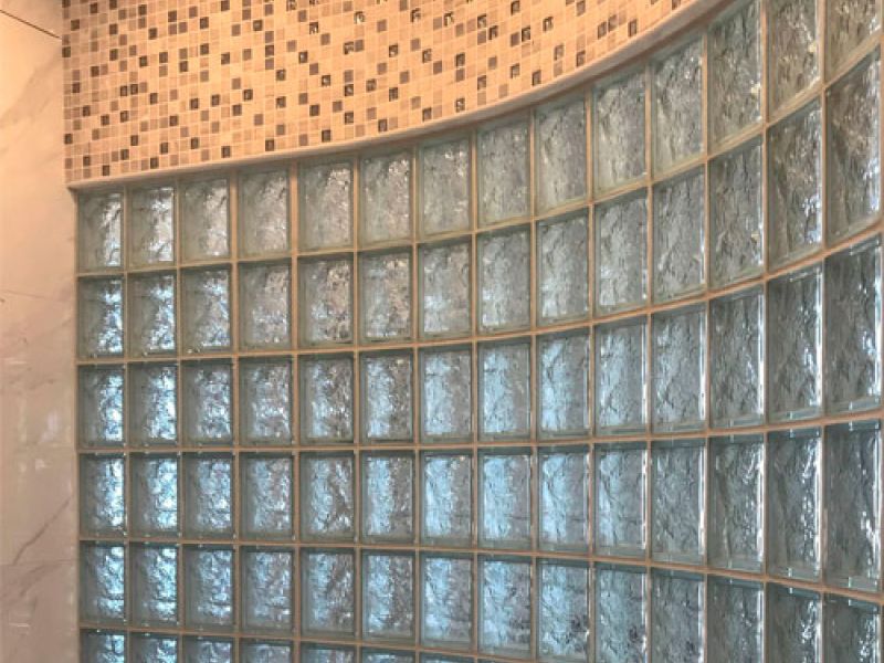 Curved glass block wall with textured glass bricks in a bathroom, beneath a mosaic tile border in beige and light brown. The wall creates a decorative and semi-transparent partition.